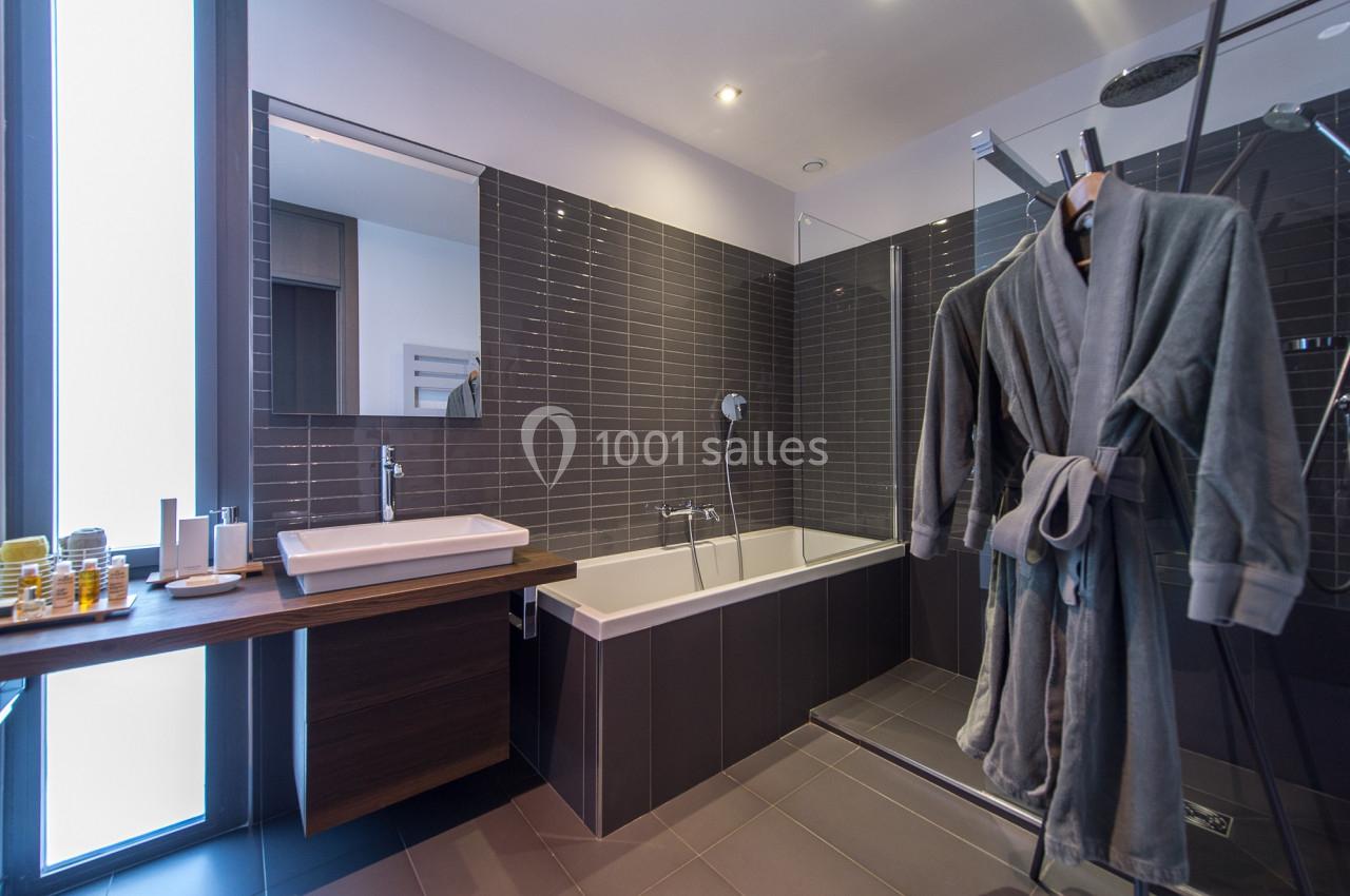 Salle de bain moderne avec baignoire, douche vitrée, lavabo et peignoirs suspendus près d'une fenêtre.