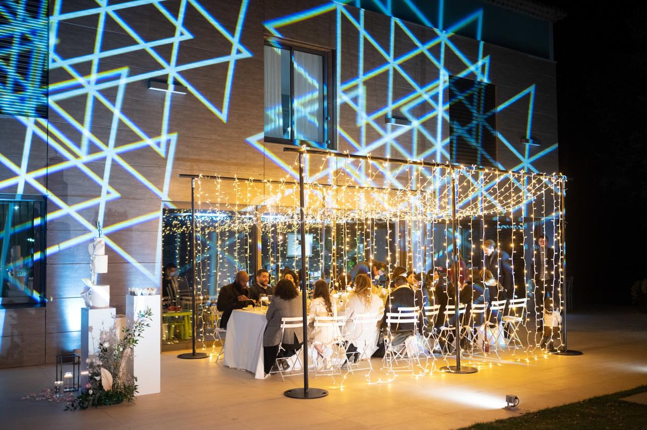 Dîner en extérieur sous une pergola illuminée de guirlandes lumineuses, avec des projections géométriques sur un mur.