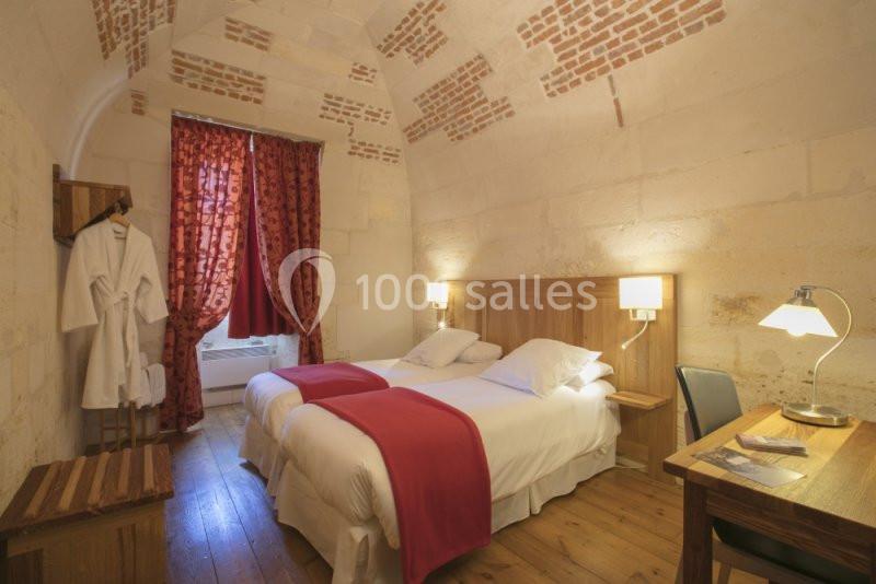 Chambre avec deux lits simples, murs en pierre, plafond voûté, rideaux rouges et éclairage chaleureux.