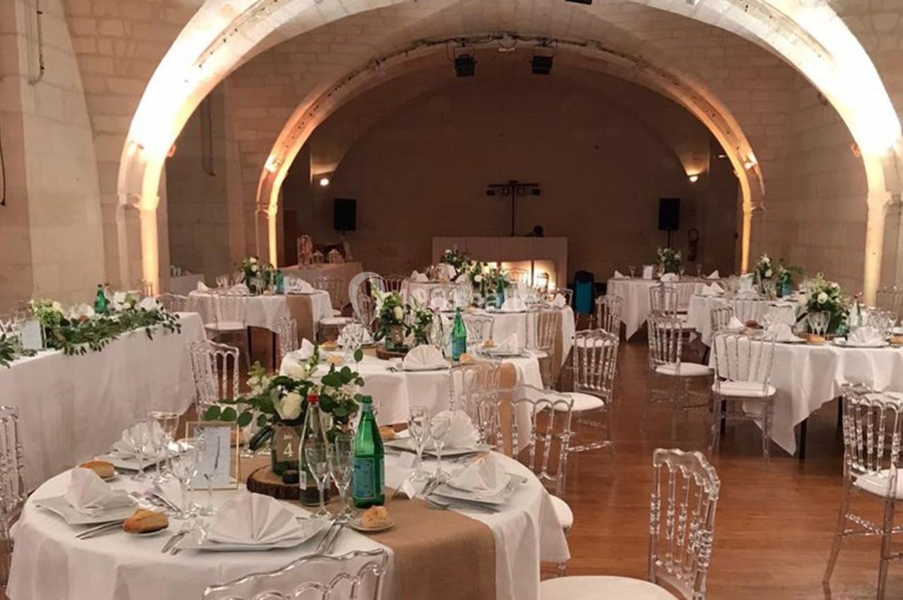 Salle voûtée décorée pour un événement, avec tables rondes dressées, nappes blanches et chaises transparentes.