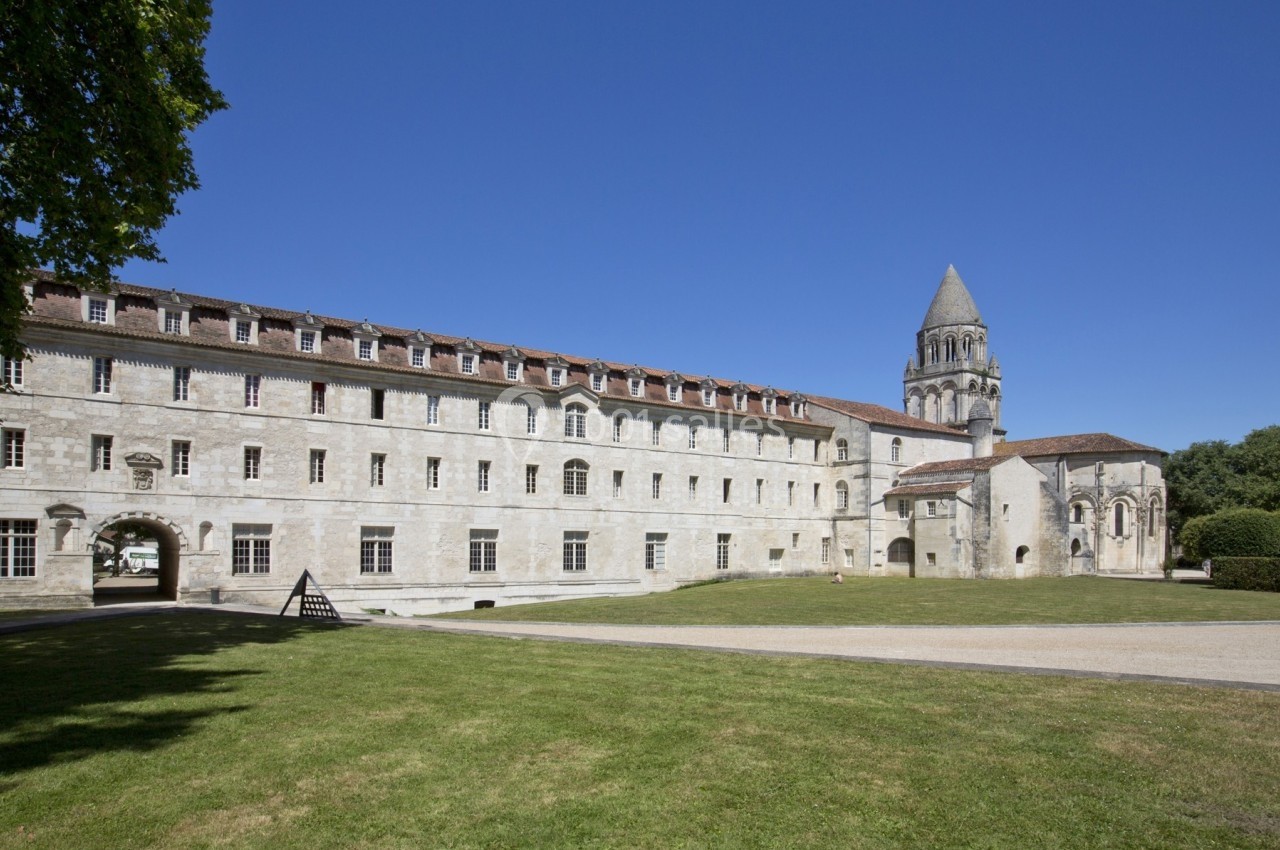 Vue d'un bâtiment historique en pierre avec une tour, entouré d'une pelouse et d'arbres sous un ciel bleu.