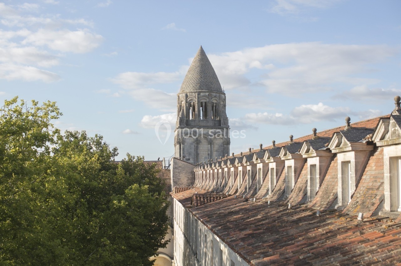 Clocher en pierre d'une église romane entouré de toits en tuiles et d'arbres sous un ciel partiellement nuageux.