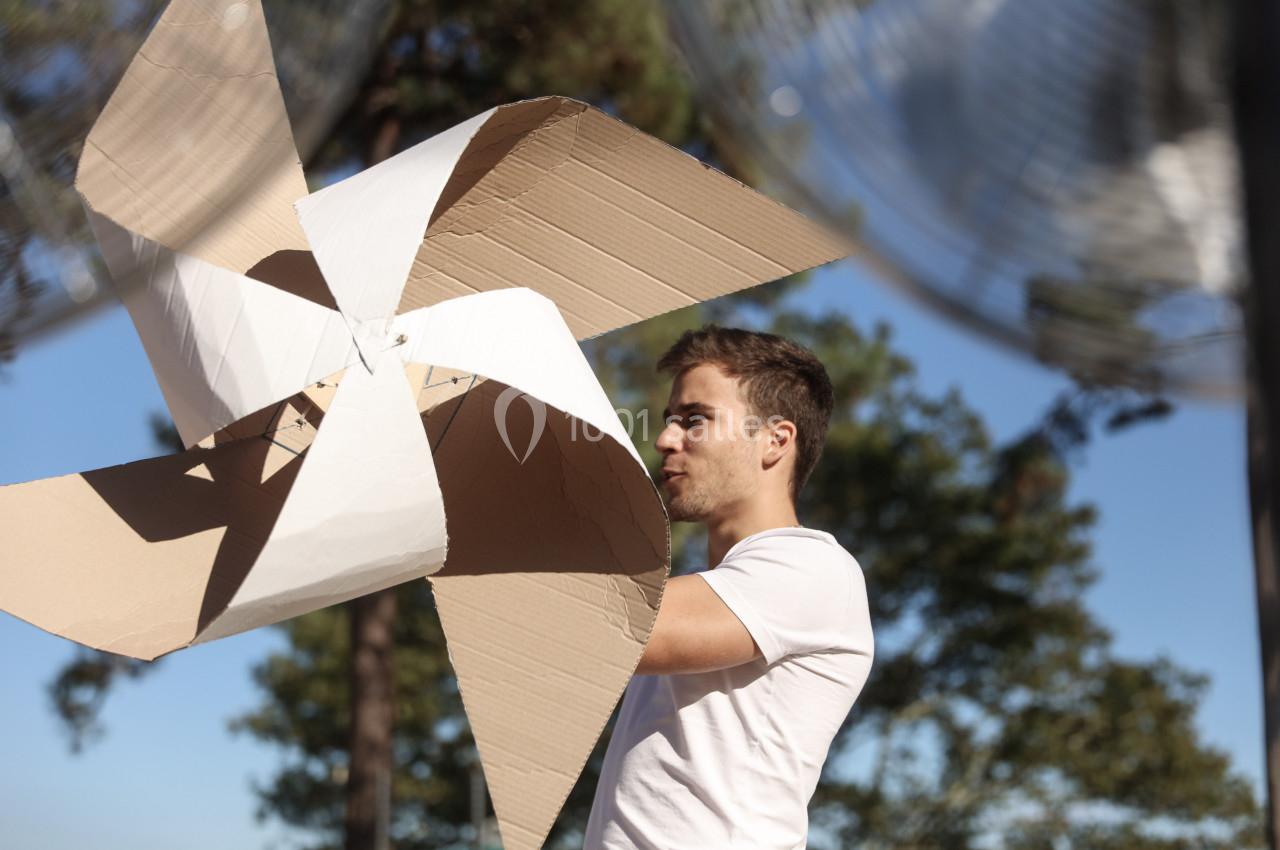 Un homme en t-shirt blanc tient une grande éolienne en carton dans un environnement extérieur ensoleillé.