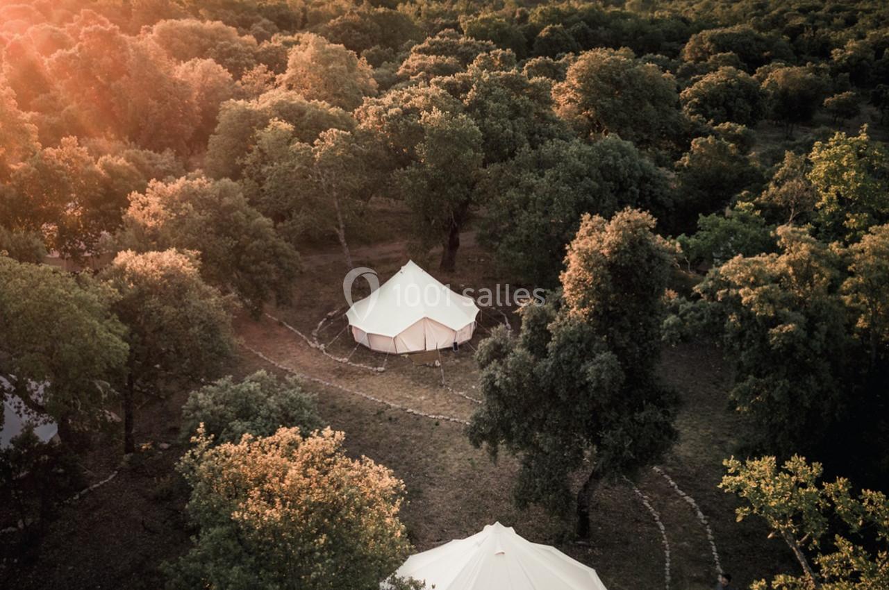 Vue aérienne de deux tentes blanches dans une clairière boisée au coucher du soleil, entourées de collines.