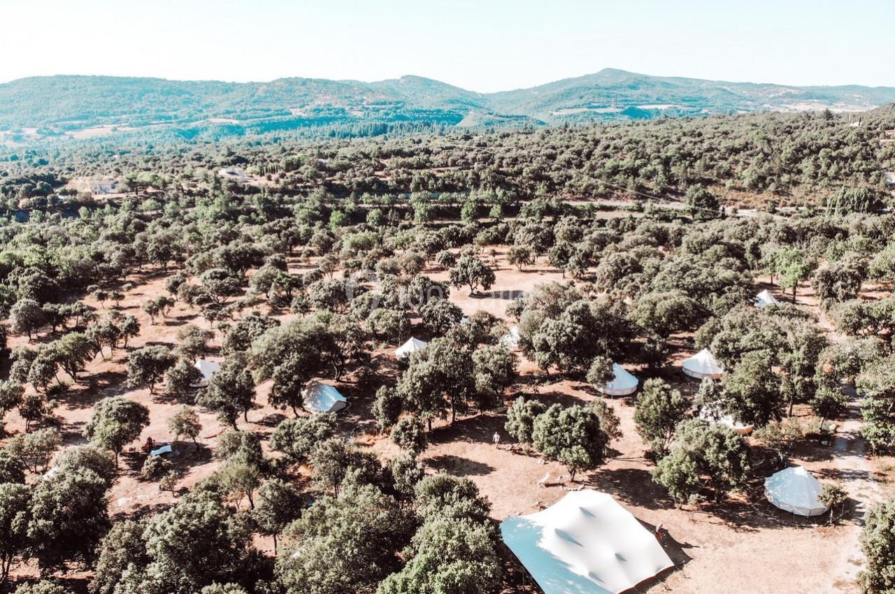 Vue aérienne d'un paysage boisé parsemé de tentes blanches, entouré de collines verdoyantes sous un ciel dégagé.