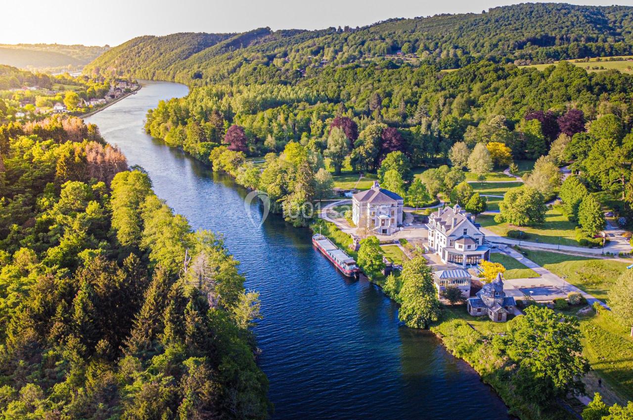 Vue aérienne d'une rivière bordée de forêts et de collines, avec un manoir et une péniche amarrée sur la rive.