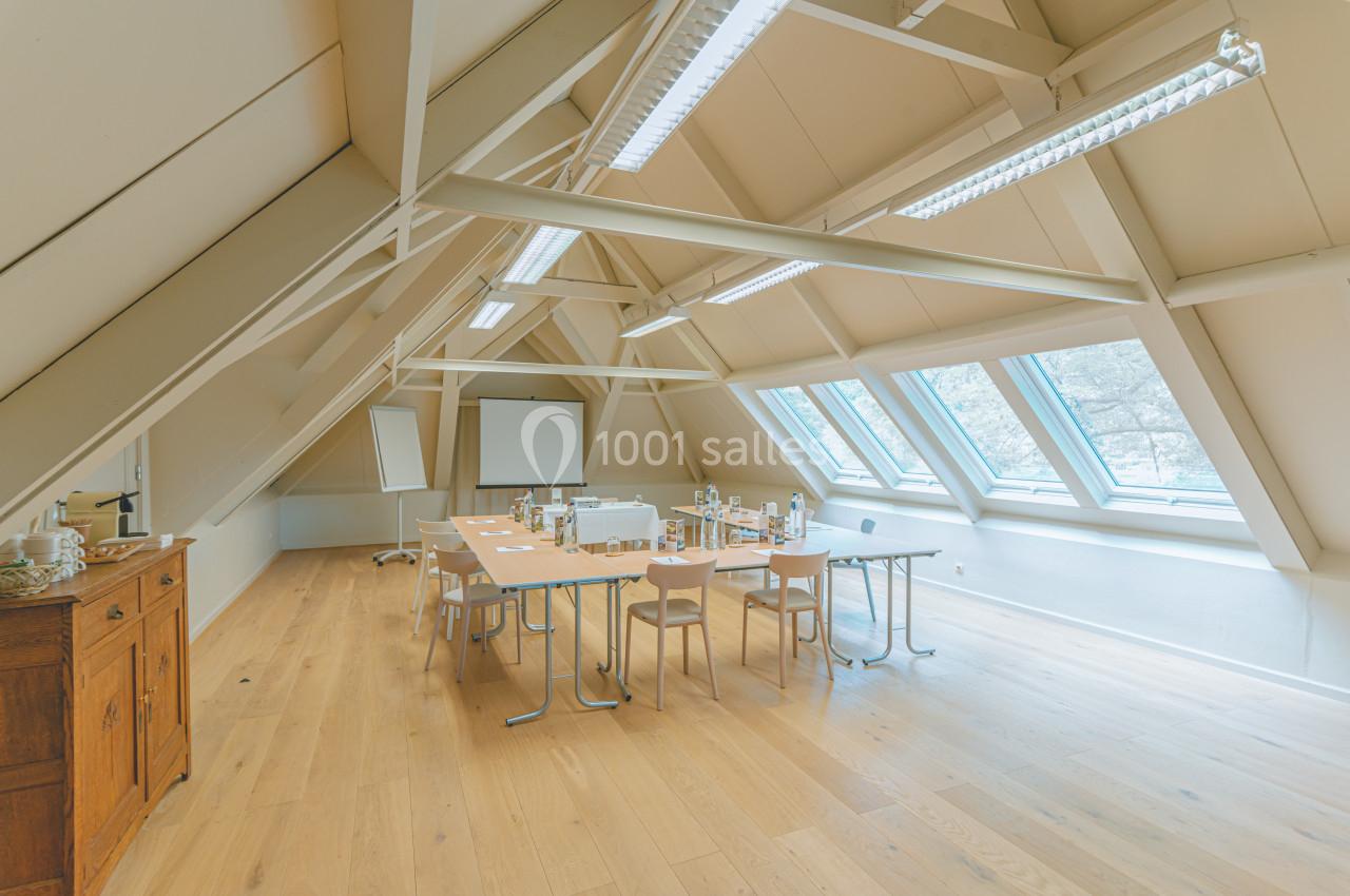 Salle de réunion lumineuse sous combles avec grandes fenêtres, table équipée de chaises et matériel de présentation.