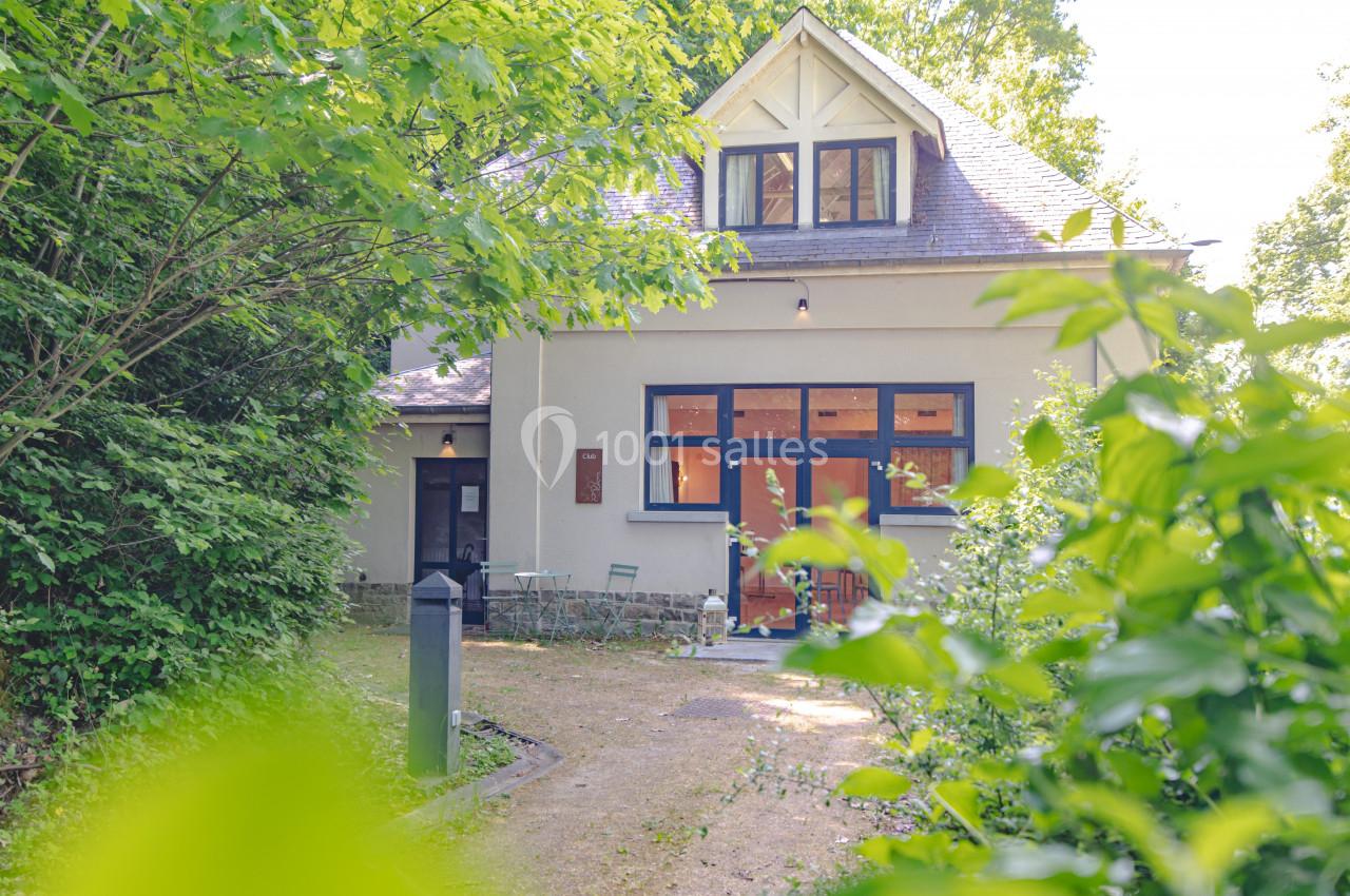 Maison entourée de verdure avec une façade claire, de grandes fenêtres et un petit espace extérieur aménagé.
