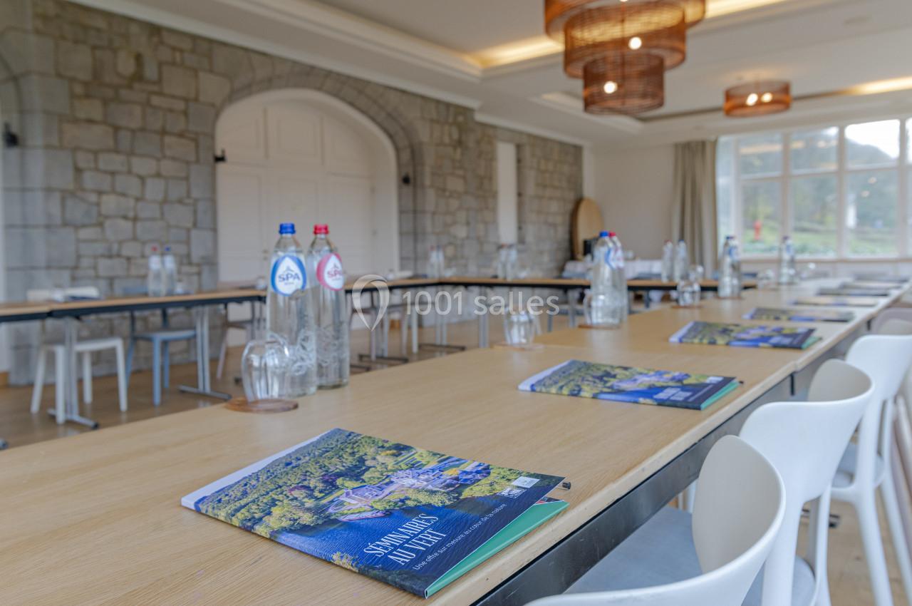 Salle de réunion lumineuse avec tables alignées, chaises blanches, bouteilles d'eau et brochures posées sur les tables.