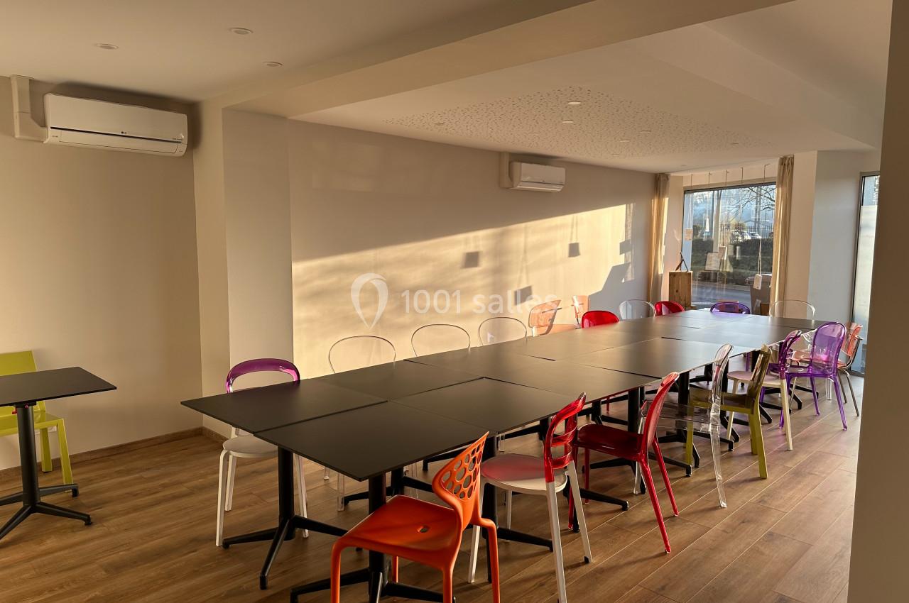 Salle lumineuse avec de grandes tables noires, chaises colorées et parquet en bois, éclairée par la lumière du soleil.