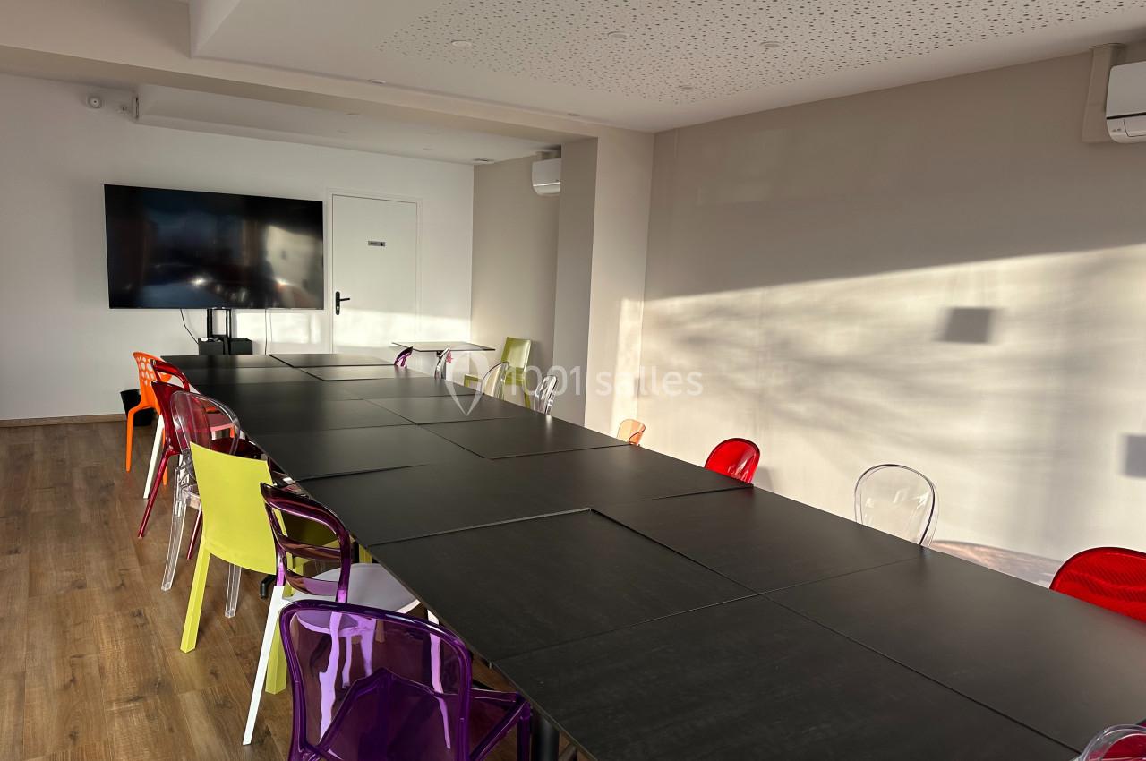 Salle de réunion lumineuse avec une grande table noire entourée de chaises colorées et un écran mural.