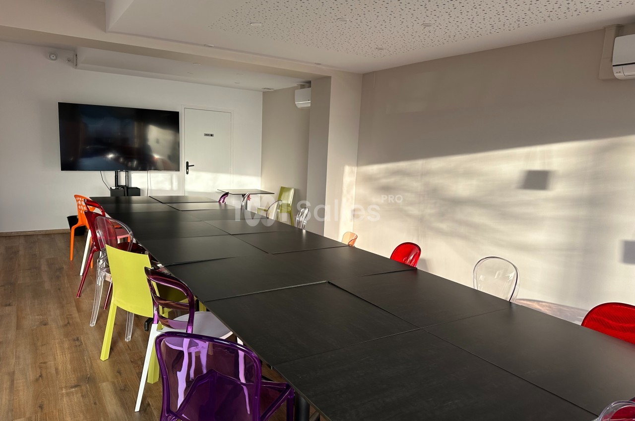 Salle de réunion lumineuse avec une grande table noire entourée de chaises colorées et un écran mural.