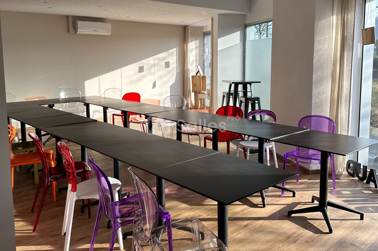 Salle lumineuse avec de longues tables noires et des chaises colorées en plastique transparent, disposées pour une réunion.