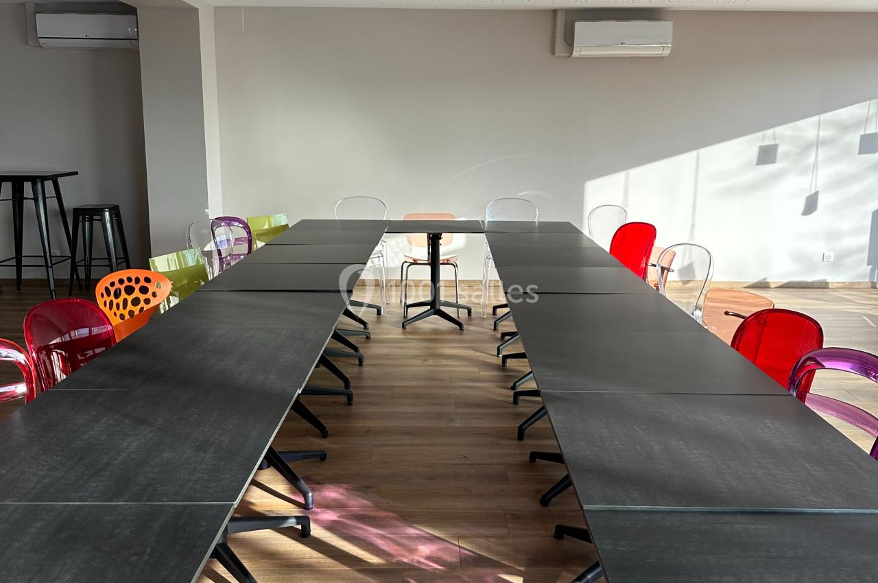 Salle de réunion lumineuse avec des tables noires disposées en U et des chaises colorées sur un sol en bois.