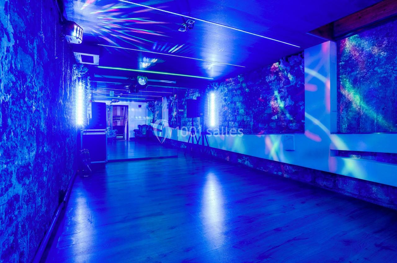 Salle avec murs en pierre, éclairage bleu et jeux de lumière colorés, sol en bois et ambiance festive.
