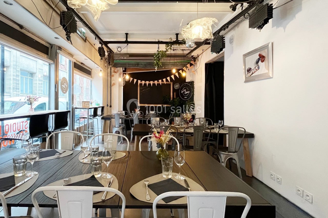 Salle de restaurant lumineuse avec tables dressées, chaises en métal, guirlande lumineuse et décoration végétale.