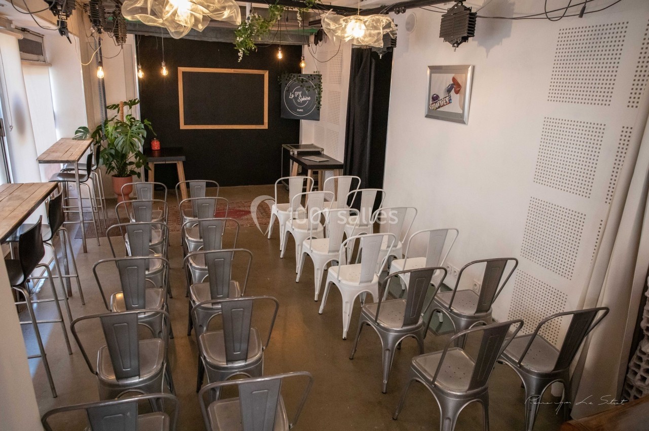 Salle aménagée avec des chaises métalliques face à un tableau noir, éclairée par des suspensions lumineuses.