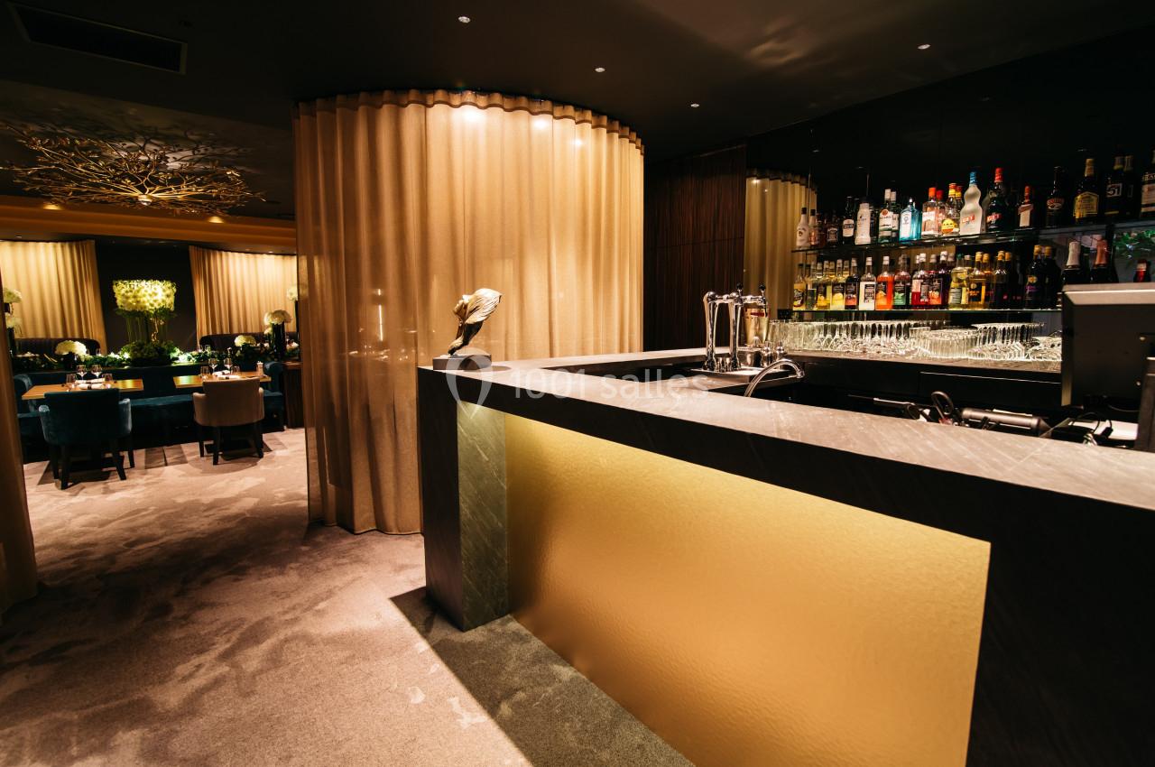 Bar élégant avec comptoir éclairé, étagères de bouteilles en arrière-plan et salle à manger visible derrière un rideau.