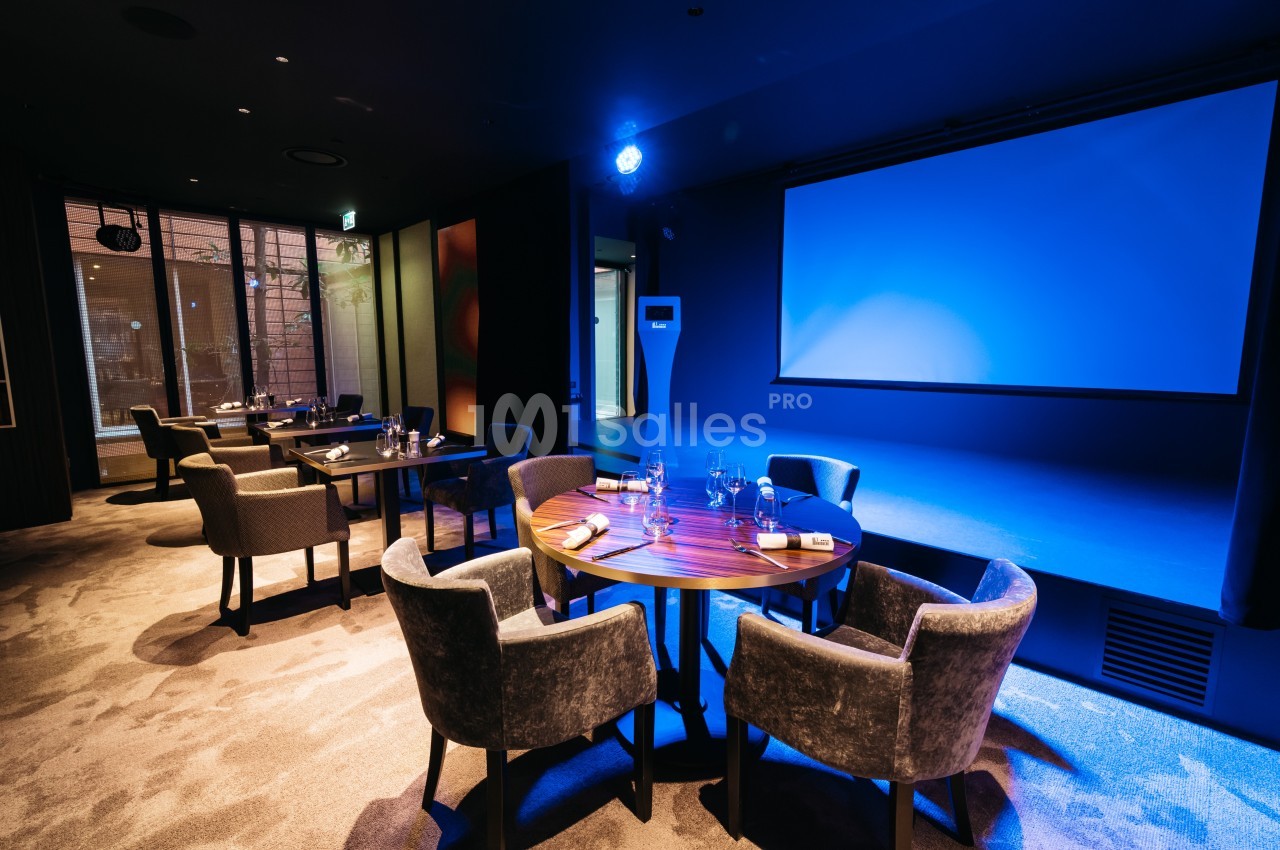 Salle de restaurant moderne avec tables rondes dressées, éclairage tamisé et écran de projection allumé en arrière-plan.