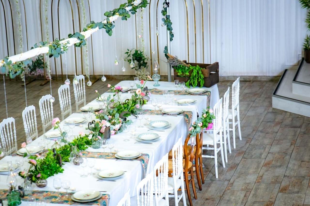 Table décorée avec nappes blanches, vaisselle, fleurs et feuillages, entourée de chaises dans une salle lumineuse.