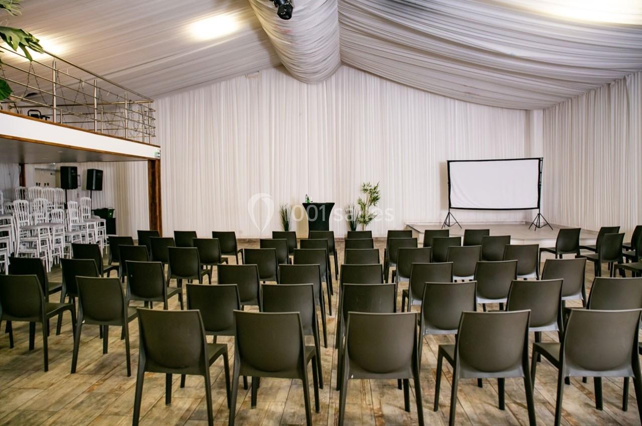 Salle de conférence avec des rangées de chaises noires, un pupitre central et un écran de projection à l'arrière.