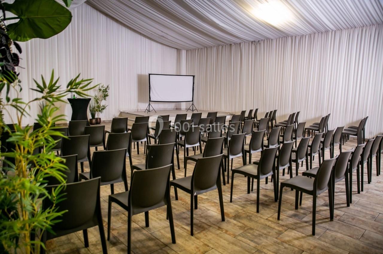 Salle de conférence avec des rangées de chaises noires alignées face à un écran de projection blanc.