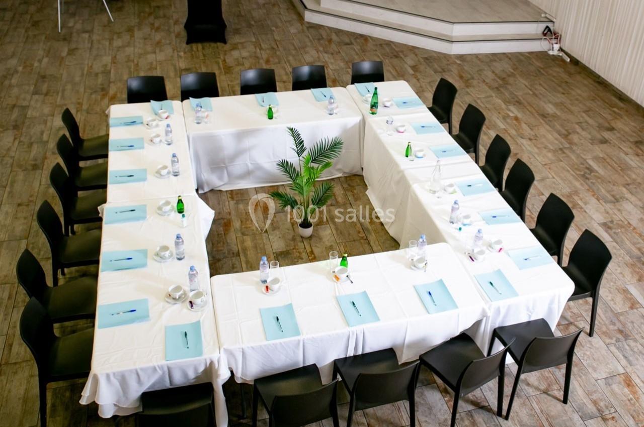 Salle de réunion avec tables disposées en U, nappes blanches, chaises noires et documents posés devant chaque place.