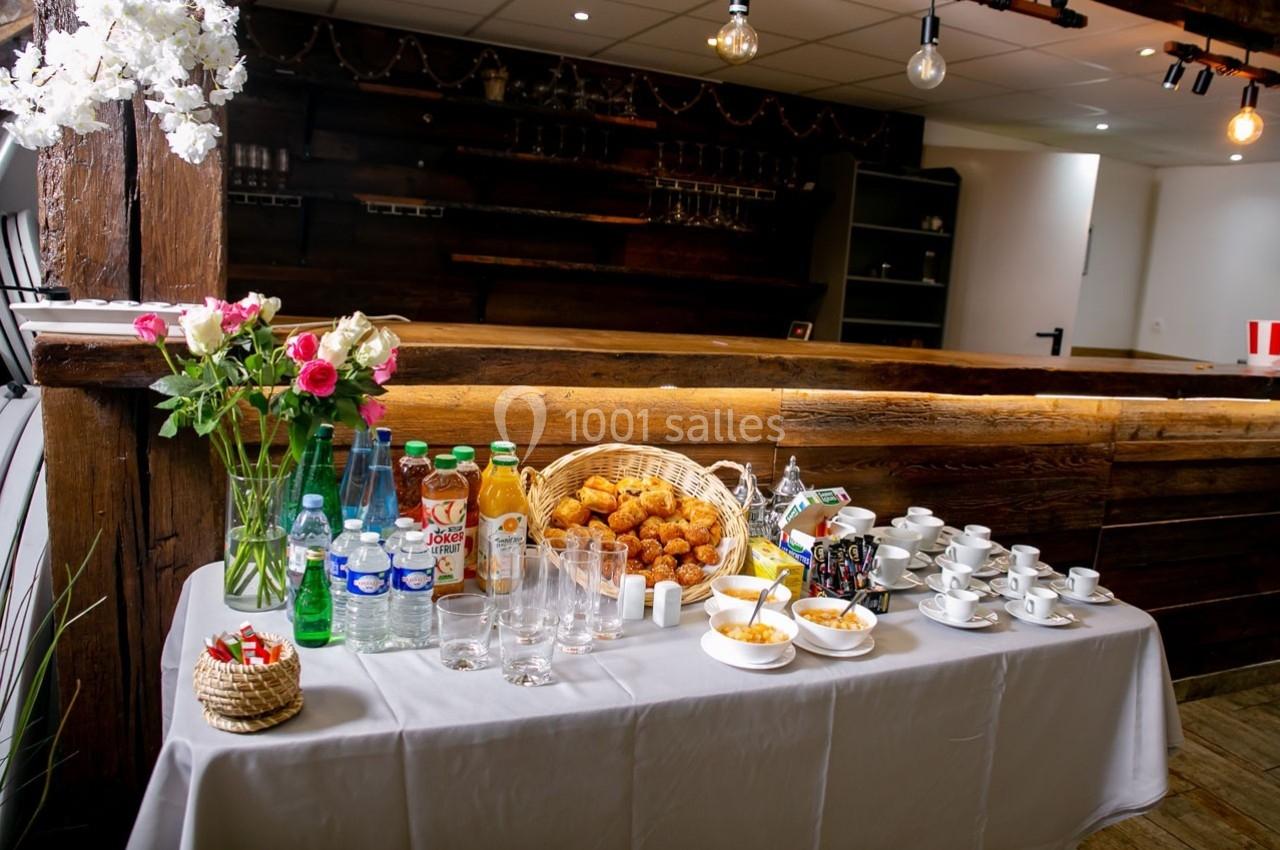 Table dressée avec boissons, viennoiseries, tasses et verres, dans un espace intérieur en bois avec éclairage chaleureux.