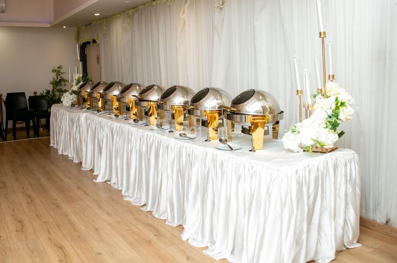 Buffet avec plusieurs plats en chafing dishes alignés sur une table drapée de blanc, décorée de fleurs.