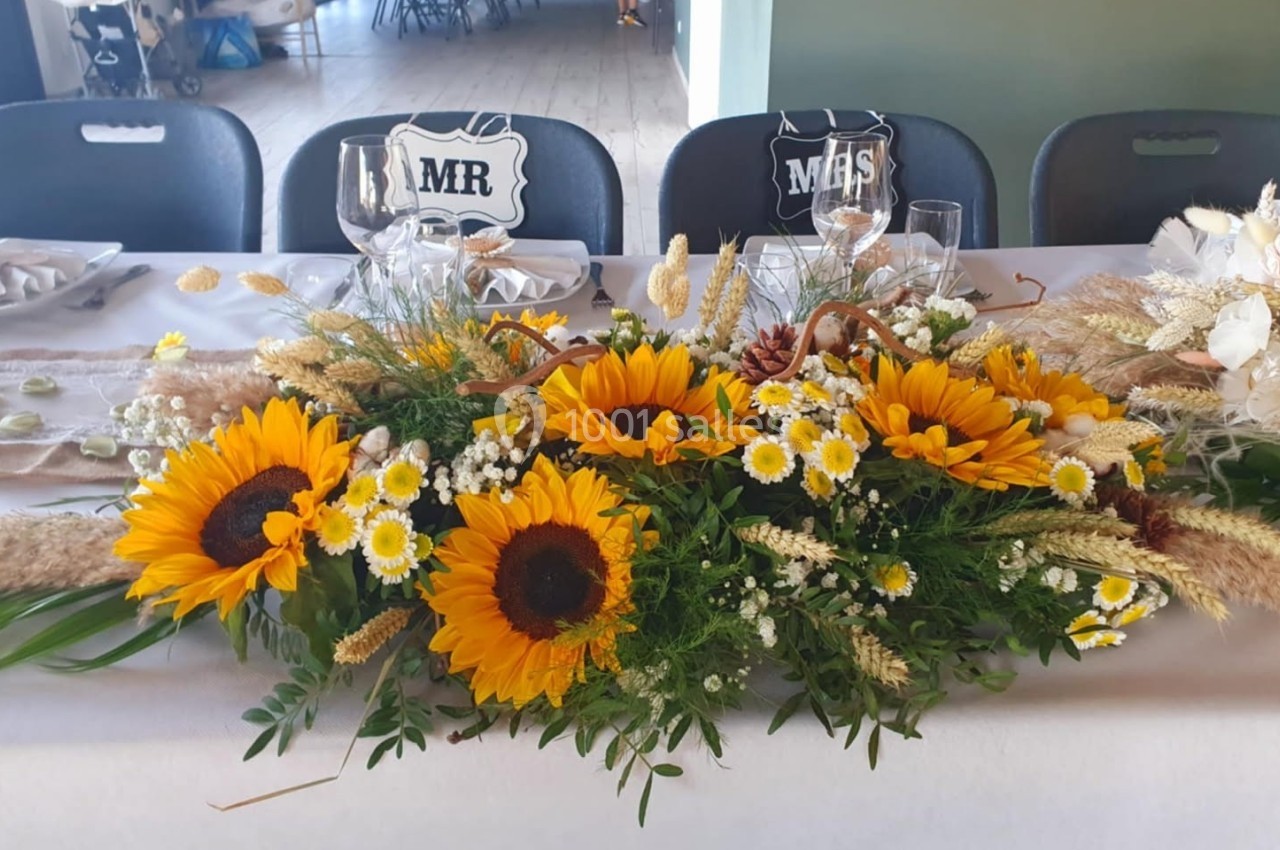 Centre de table avec des tournesols, des marguerites et des herbes séchées, disposé sur une table de réception.