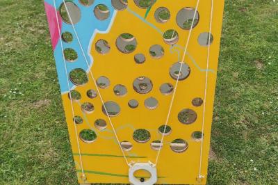 Planche de jeu de lancer de sacs colorée posée sur une pelouse, avec des sacs verts et jaunes dispersés autour.