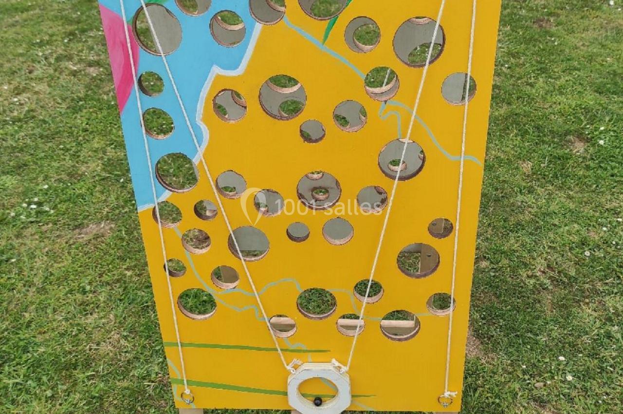 Panneau en bois jaune percé de trous, représentant un fromage, utilisé pour un jeu en plein air sur une pelouse.