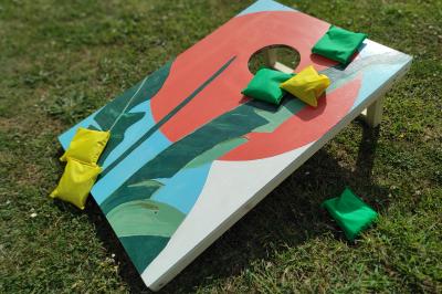 Planche de jeu de lancer de sacs colorée posée sur une pelouse, avec des sacs verts et jaunes dispersés autour.