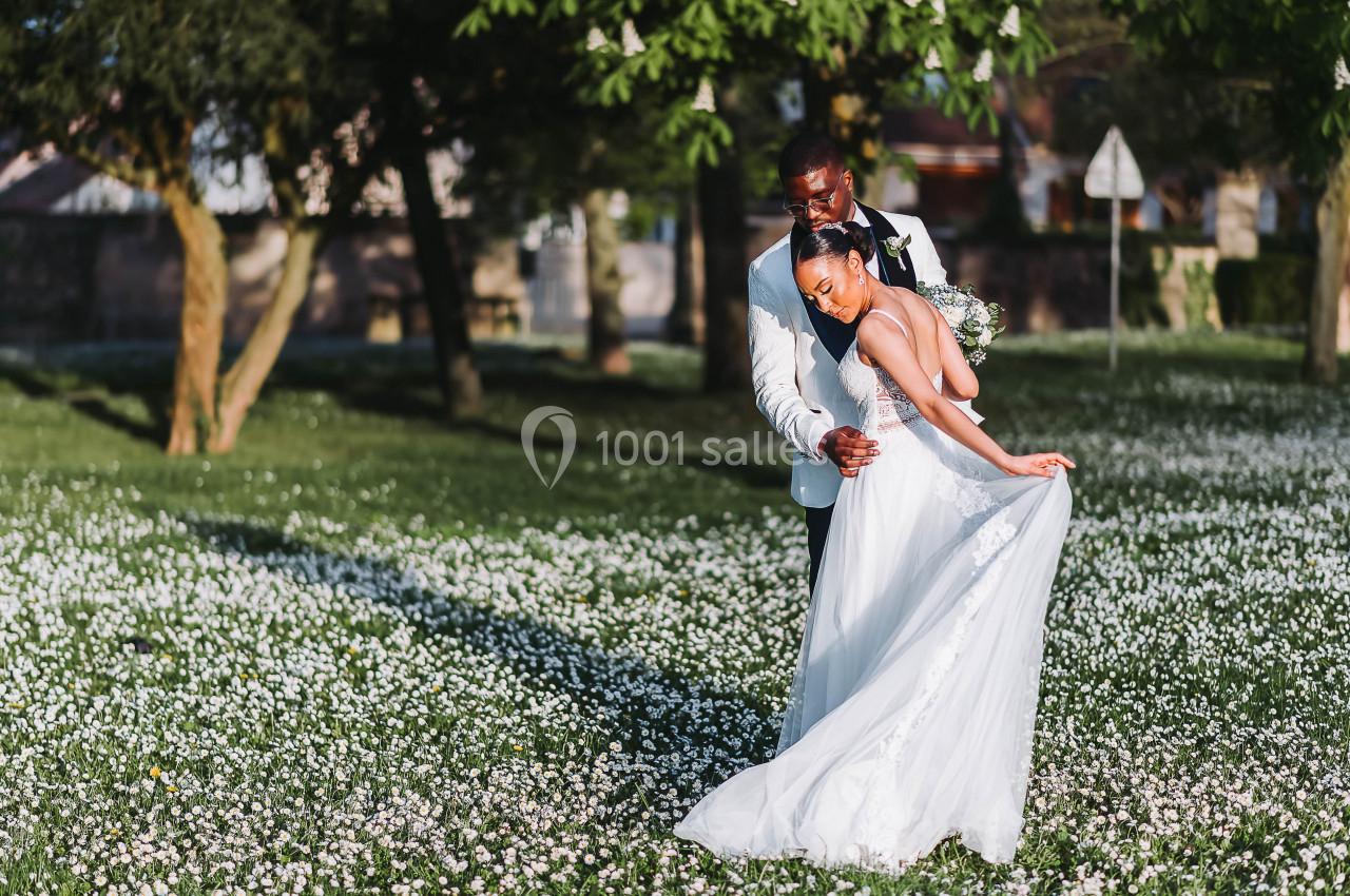 Un couple en tenue de mariage pose dans un champ fleuri sous des arbres par une journée ensoleillée.