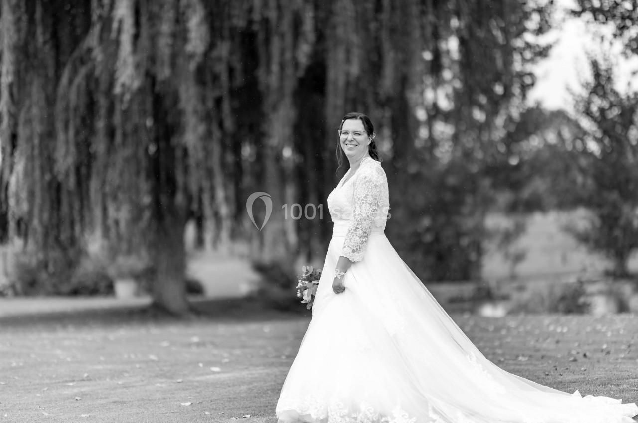 Femme en robe de mariée blanche tenant un bouquet, debout sur une pelouse devant un grand saule pleureur.