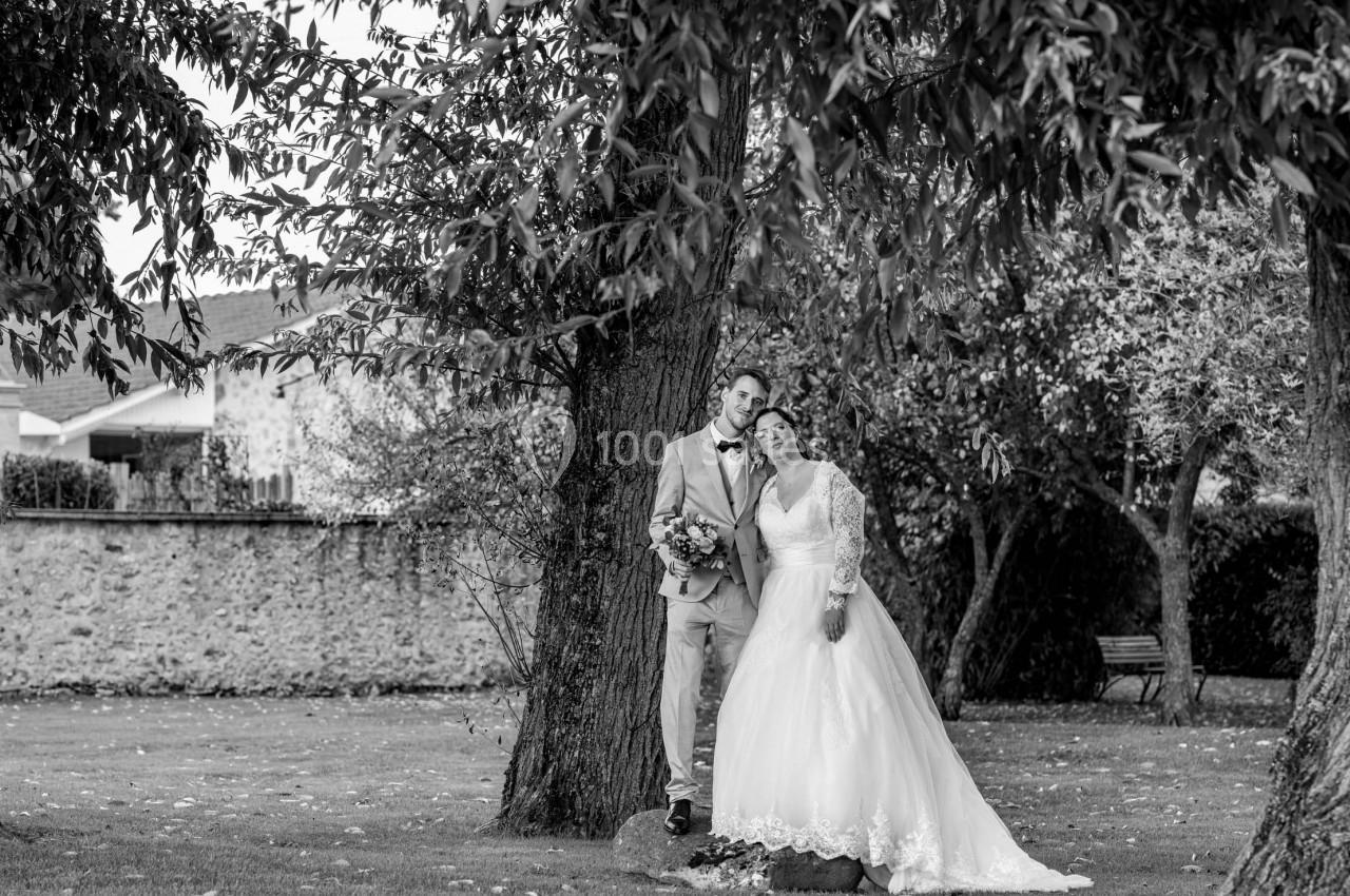 Un couple en tenue de mariage pose sous des arbres dans un jardin, avec un mur en pierre en arrière-plan.