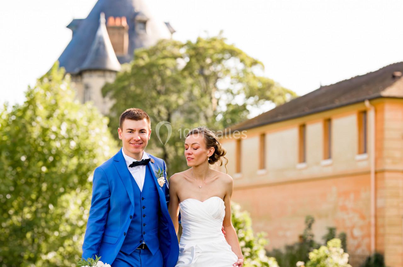 Un couple en tenue de mariage marche dans un jardin, avec un bâtiment et des arbres en arrière-plan.