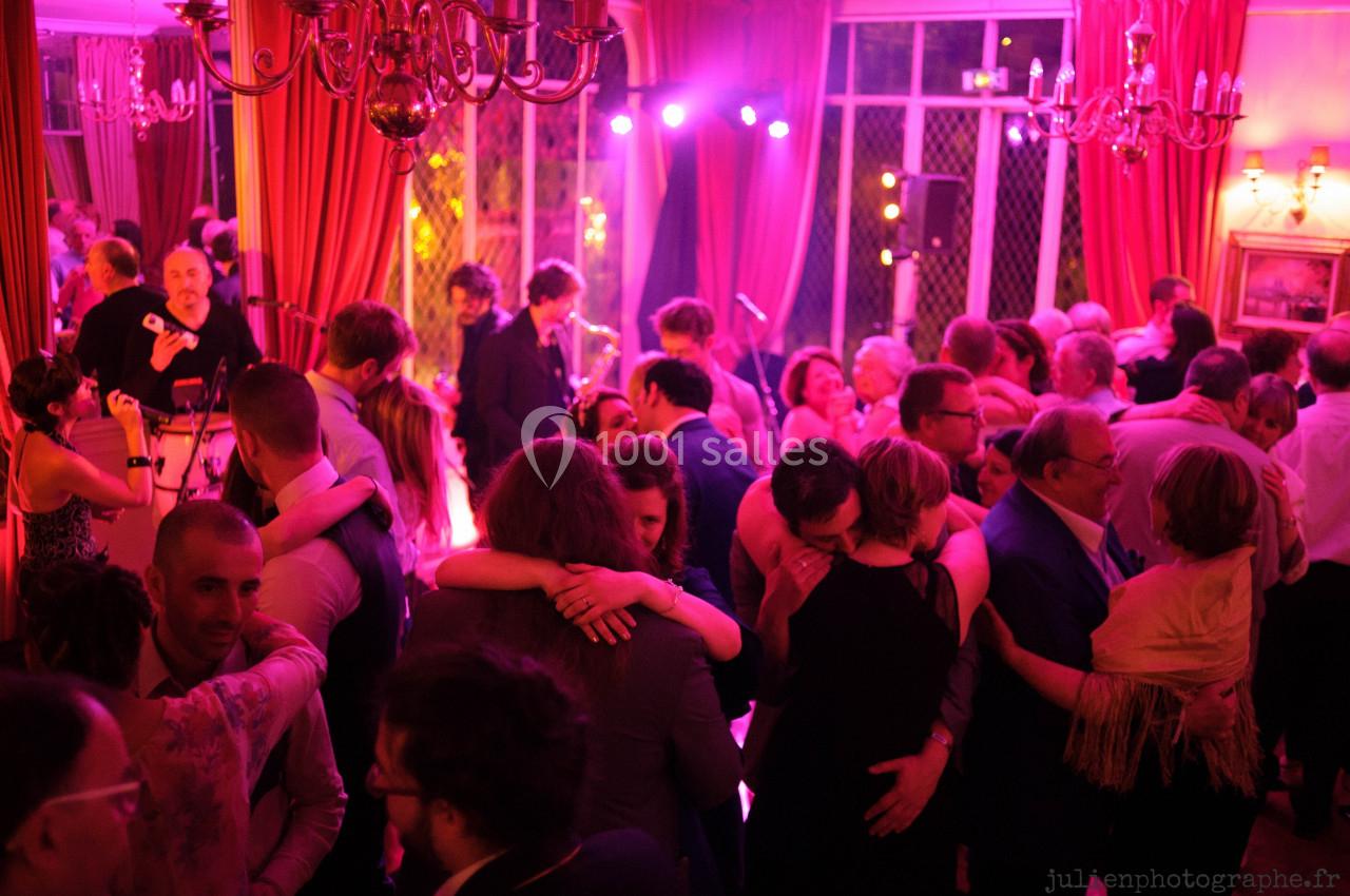 Des personnes dansent et discutent dans une salle éclairée par des lumières colorées, avec un groupe de musique en arrière…