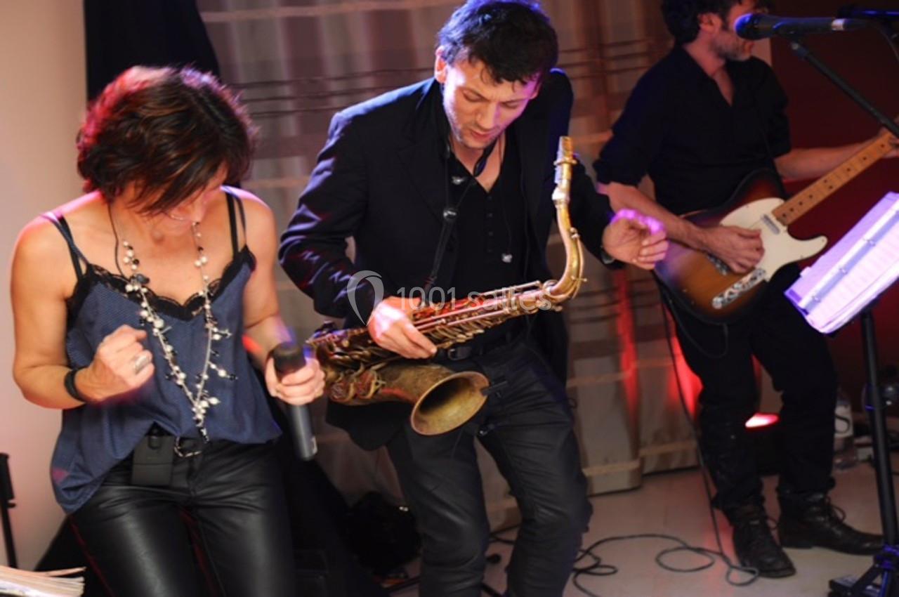 Un groupe de musiciens joue sur scène, une femme chante tandis qu'un homme joue du saxophone et un autre de la guitare.