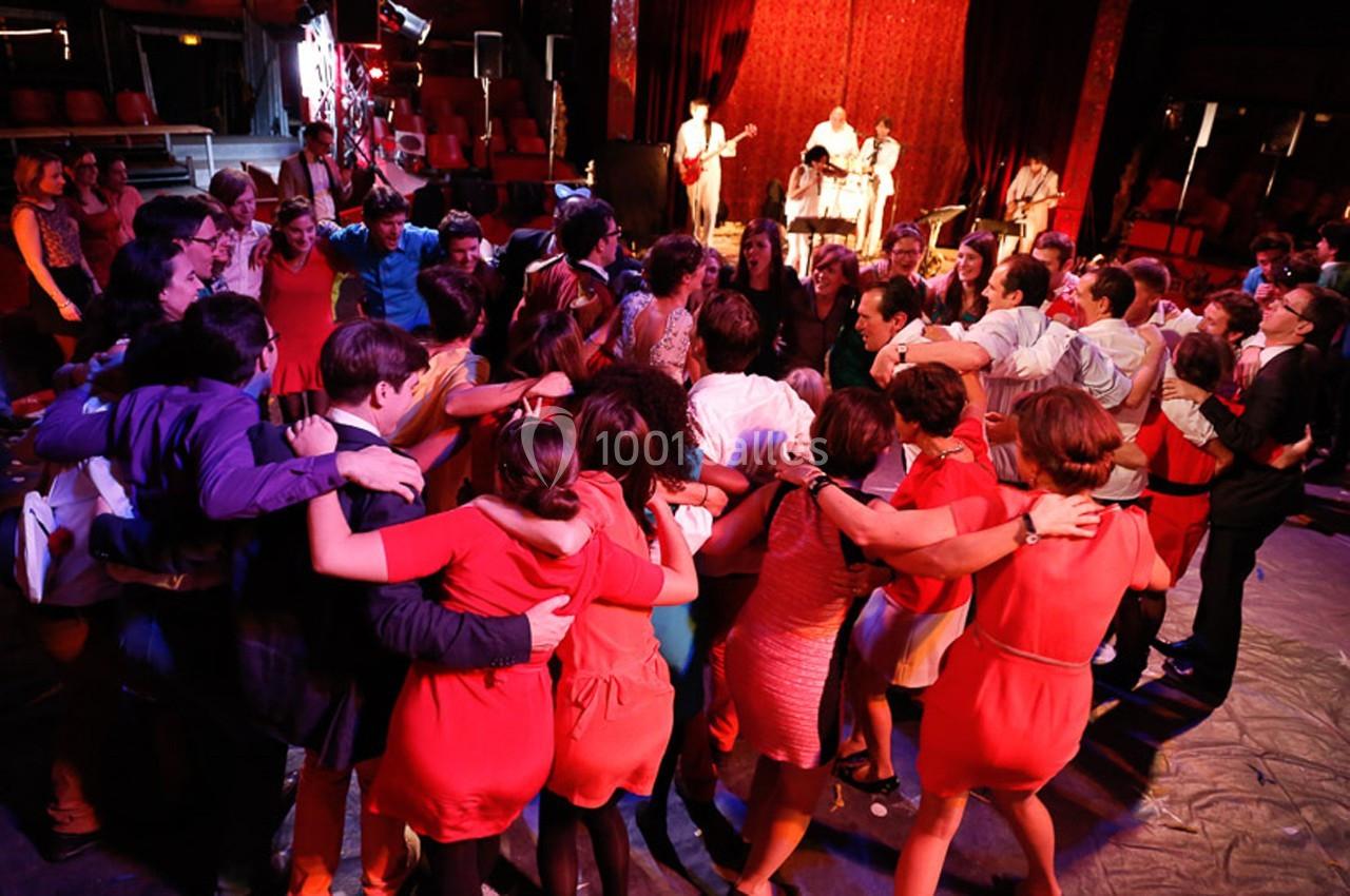 Un groupe de personnes dansant en cercle dans une salle éclairée avec un groupe de musiciens en arrière-plan.