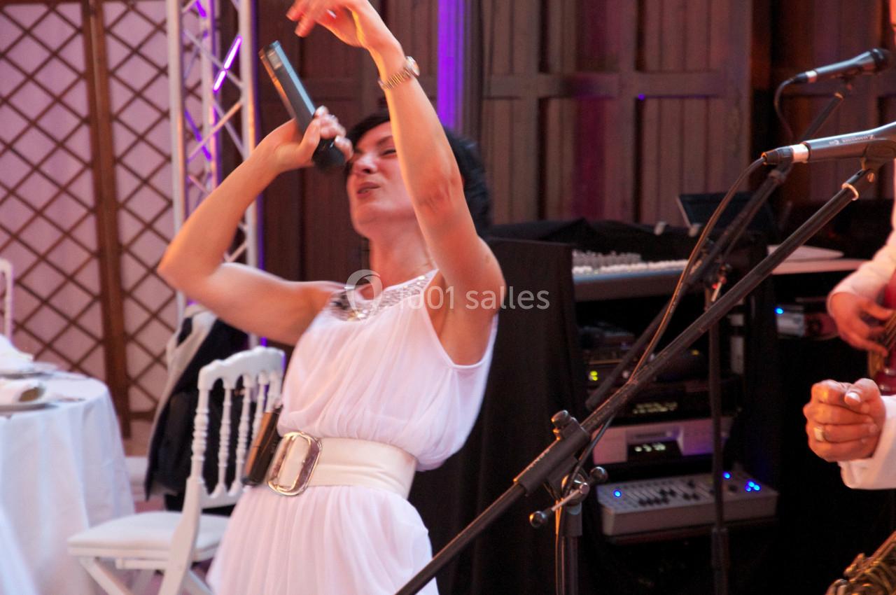 Femme en robe blanche chantant sur scène avec un microphone, entourée d'équipements audio et d'instruments.