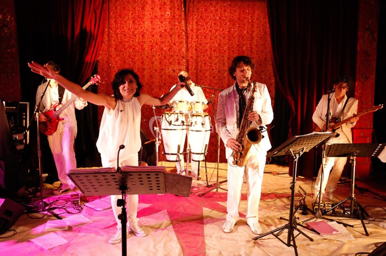 Groupe de musiciens en tenue blanche jouant sur scène avec éclairage rouge et instruments variés.