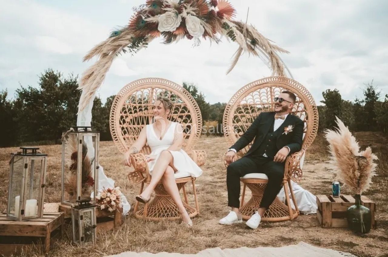 Location salle Saint-Genès-de-Lombaud (Gironde) - Domaine du Château Icla #2 Un couple assis sur des fauteuils en rotin sous une arche décorée de fleurs séchées dans un cadre extérieur champêtre.