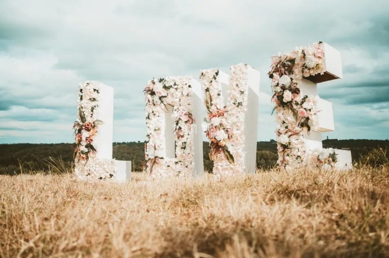Location salle Saint-Genès-de-Lombaud (Gironde) - Domaine du Château Icla #9 Lettres géantes formant le mot ’LOVE’, décorées de fleurs, placées dans un champ sous un ciel nuageux.