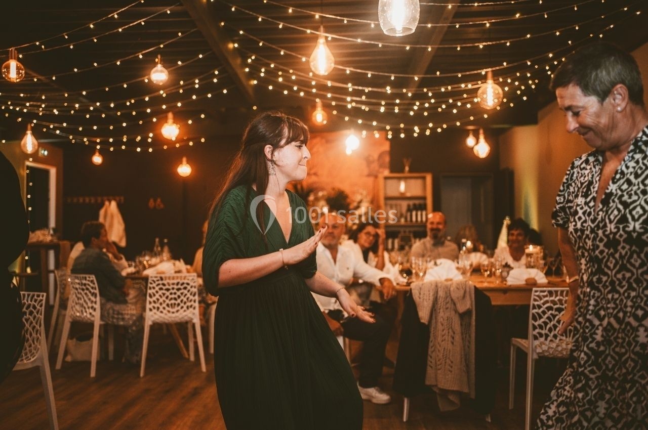 Location salle Saint-Genès-de-Lombaud (Gironde) - Domaine du Château Icla #14 Une femme danse dans une salle décorée de guirlandes lumineuses, entourée de convives assis à des tables.