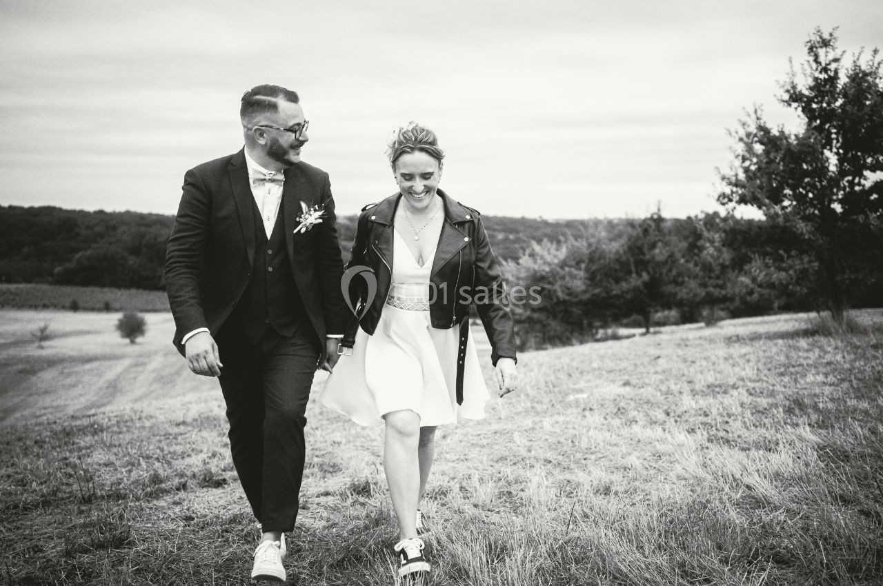 Location salle Saint-Genès-de-Lombaud (Gironde) - Domaine du Château Icla #22 Un couple souriant marche dans un champ, elle en robe et blouson, lui en costume, sous un ciel nuageux.