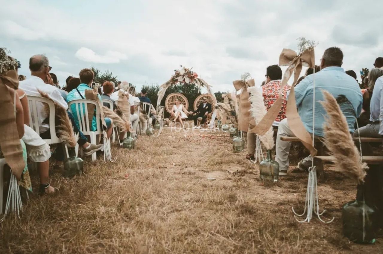 Location salle Saint-Genès-de-Lombaud (Gironde) - Domaine du Château Icla #23 Cérémonie de mariage en plein air avec des invités assis et une arche décorée au centre sous un ciel nuageux.