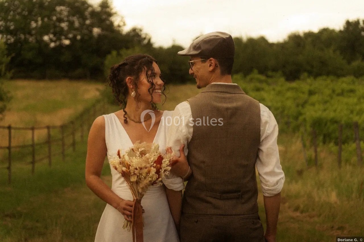Location salle Saint-Genès-de-Lombaud (Gironde) - Domaine du Château Icla #31 Un couple en tenue de mariage se tient dans un vignoble, la mariée tenant un bouquet de fleurs séchées.