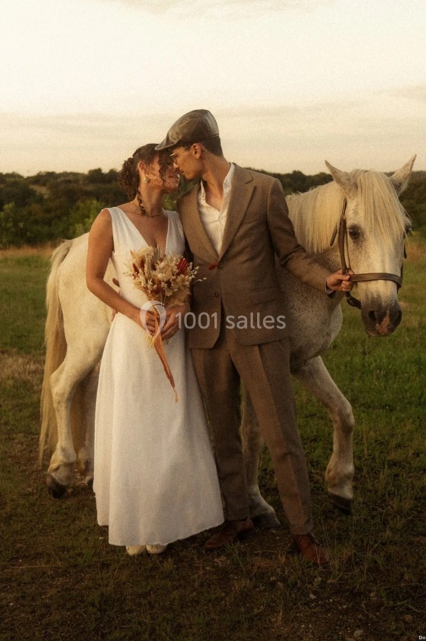 Location salle Saint-Genès-de-Lombaud (Gironde) - Domaine du Château Icla #33 Un couple en tenue de mariage pose près d'un cheval blanc dans un champ au coucher du soleil.