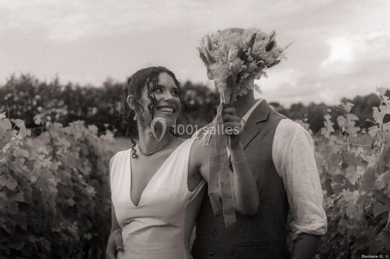 Location salle Saint-Genès-de-Lombaud (Gironde) - Domaine du Château Icla #34 Un couple dans un vignoble, la femme sourit tandis que l'homme cache son visage avec un bouquet de fleurs.